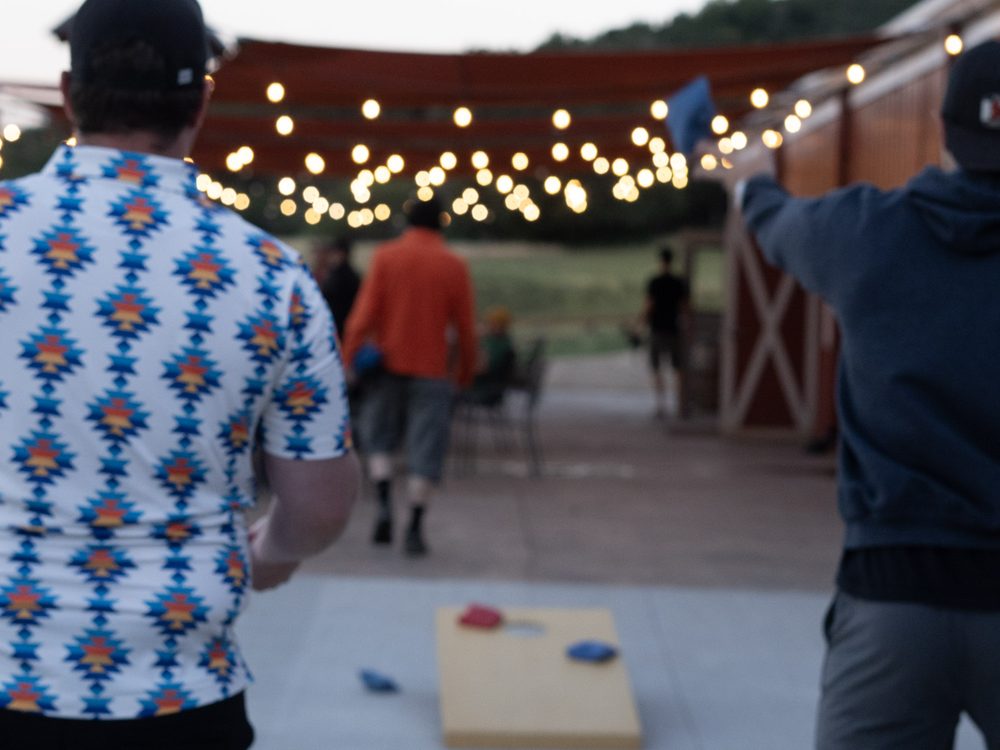 Men playing cornhole under string lights